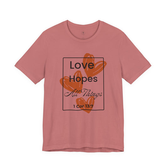 Christ Centered Faith women's Christian t-shirt with "Love Hopes All Things" and 1 Corinthians 13:7 Bible verse text in black over red hearts inside a black rectangle outline on pink background.