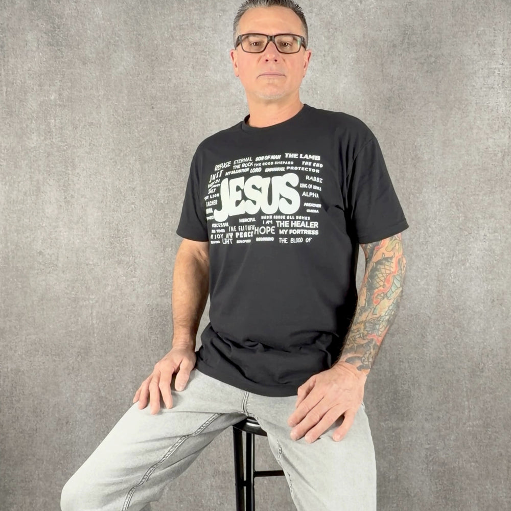 Man wearing black Christian t-shirt featuring the names of Jesus graphic design by Christ Centered Faith on a gray background.