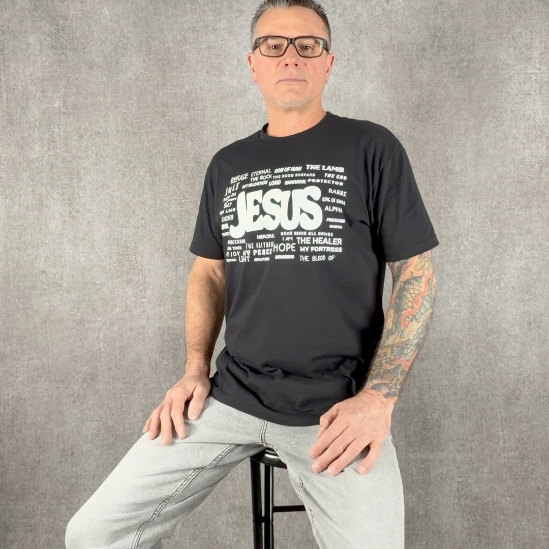 Man wearing black Christian t-shirt featuring the names of Jesus graphic design by Christ Centered Faith on a gray background.