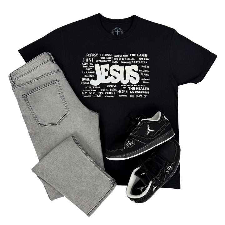 Black Christian t-shirt featuring the names of Jesus graphic designed by Christ Centered Faith styled with gray jeans and black sneakers on a white background.